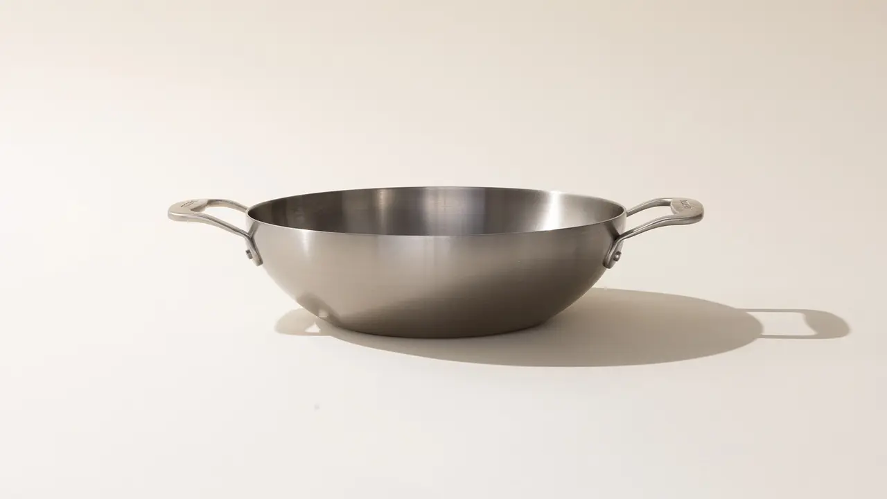 A stainless steel wok with two handles on a neutral background, casting a shadow.