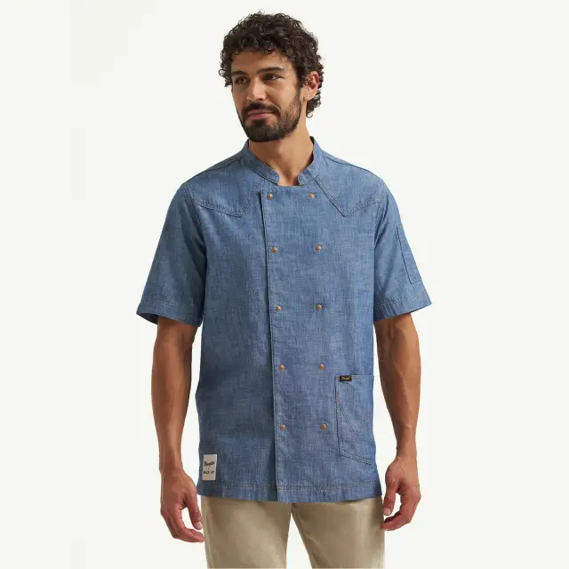 A man with curly hair wears a short-sleeve, denim-style chef's jacket and khaki pants, posing against a plain background.