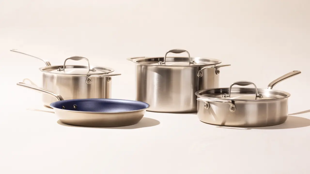 A set of stainless steel cookware including two pots with lids, a saucepan, and a skillet with a blue interior, displayed against a neutral background.