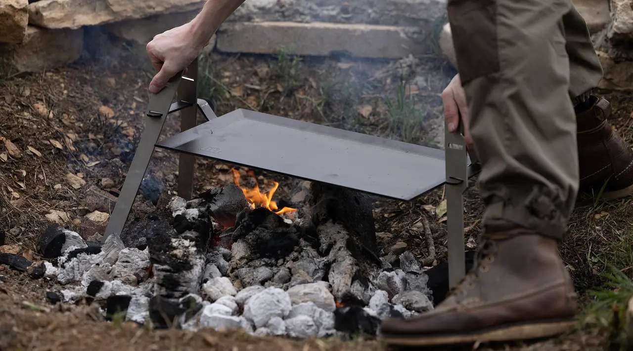A person is using a portable metal grill over an open fire outdoors.