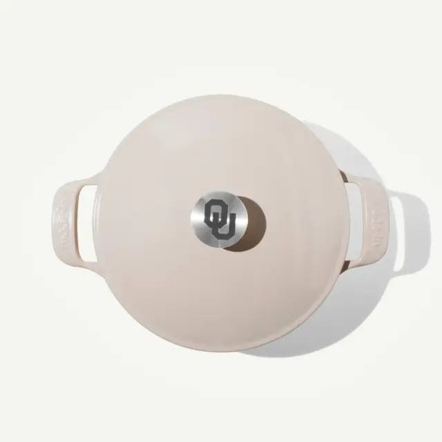 A light-colored Dutch oven with a metallic lid featuring a stylized "OU" logo is displayed from above.