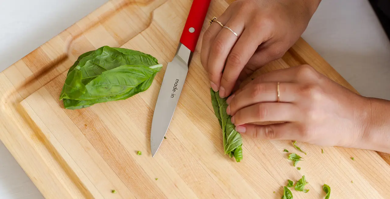 Hands are shown chopping fresh basil on a wooden cutting board with a red-handled kitchen knife.