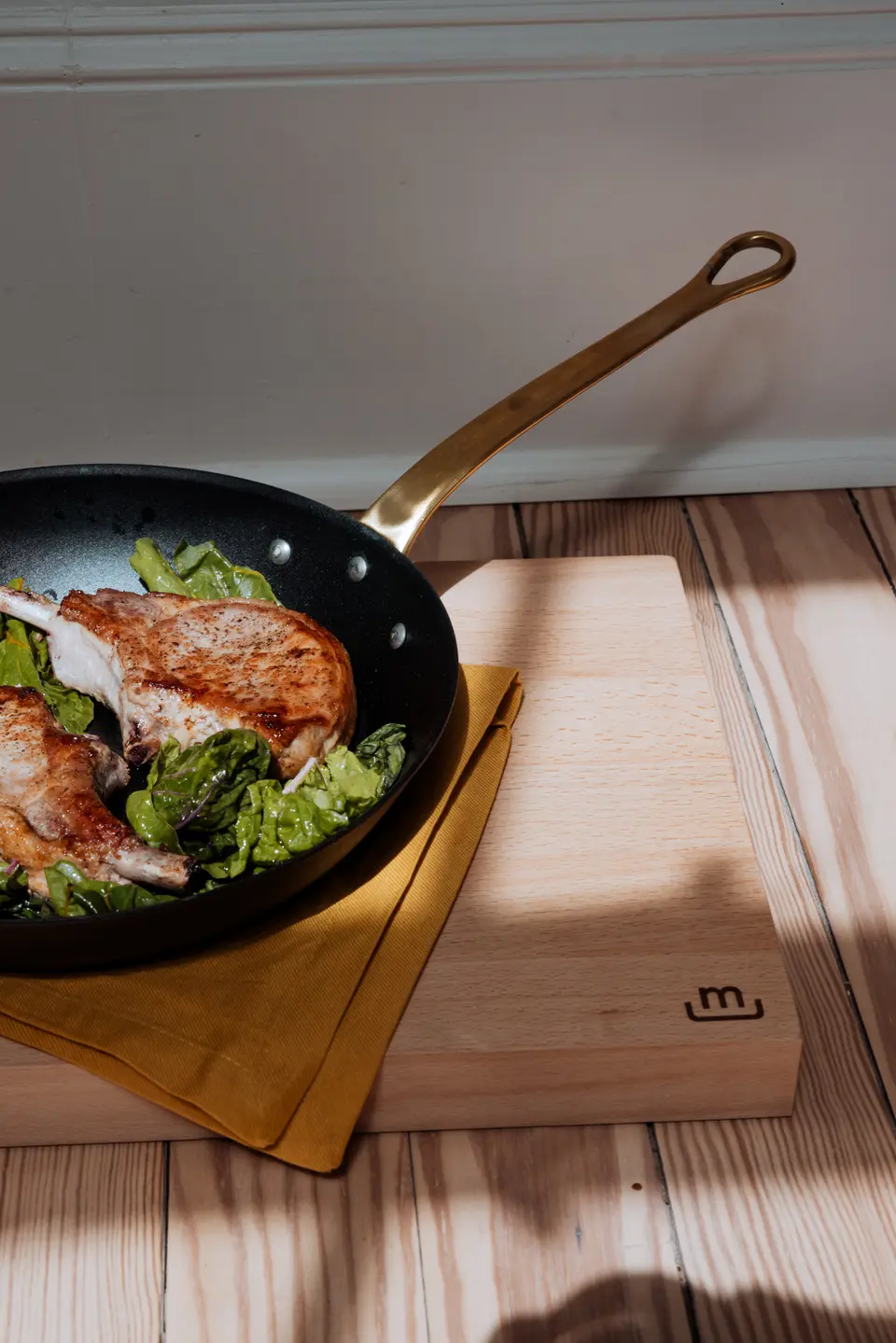A skillet with seared chicken on a bed of greens is placed on a wooden cutting board on a countertop, bathed in sunlight.