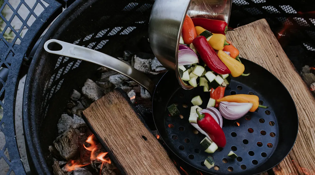 A variety of colorful vegetables are being poured into a black skillet on a wood-fired grill.