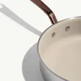 A sleek, silver cooking pot with a smooth white interior and a curved handle cast shadows on a light surface.