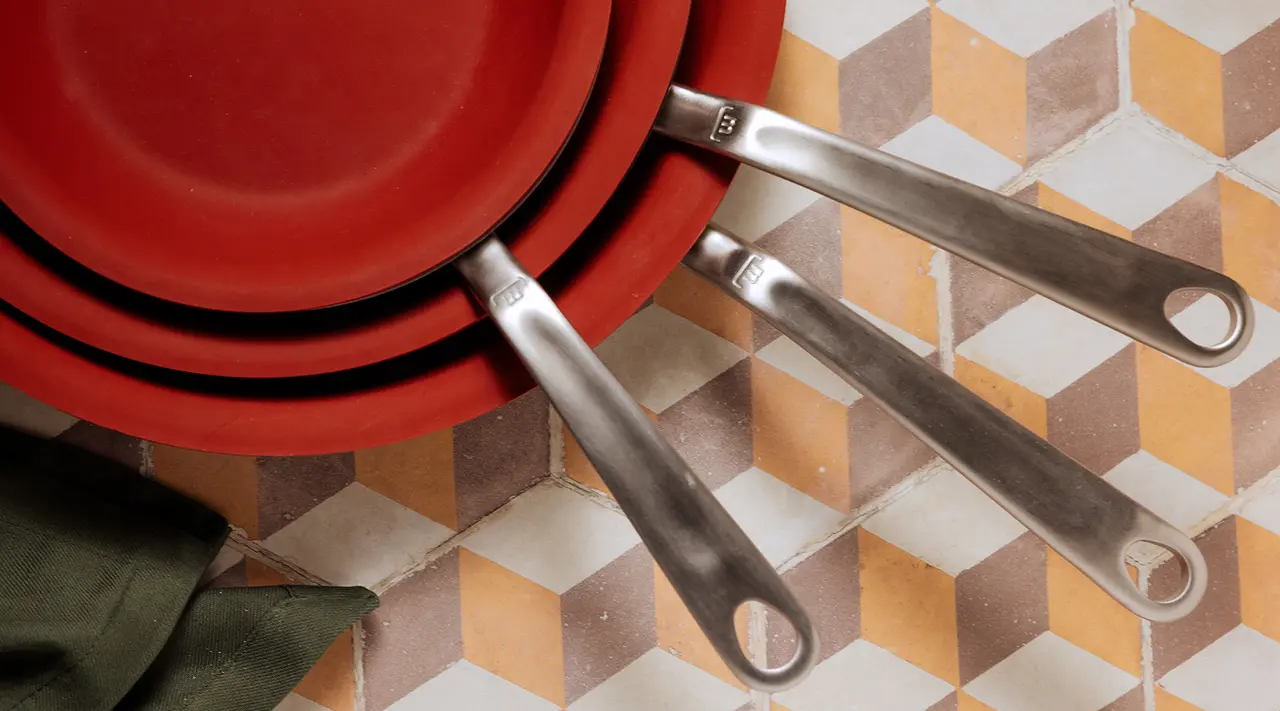 A stack of red pans with silver handles lies on a tiled surface next to a folded green cloth.