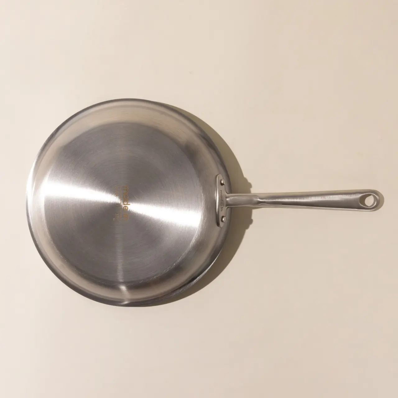 A stainless steel skillet with a long handle viewed from above on a beige background.