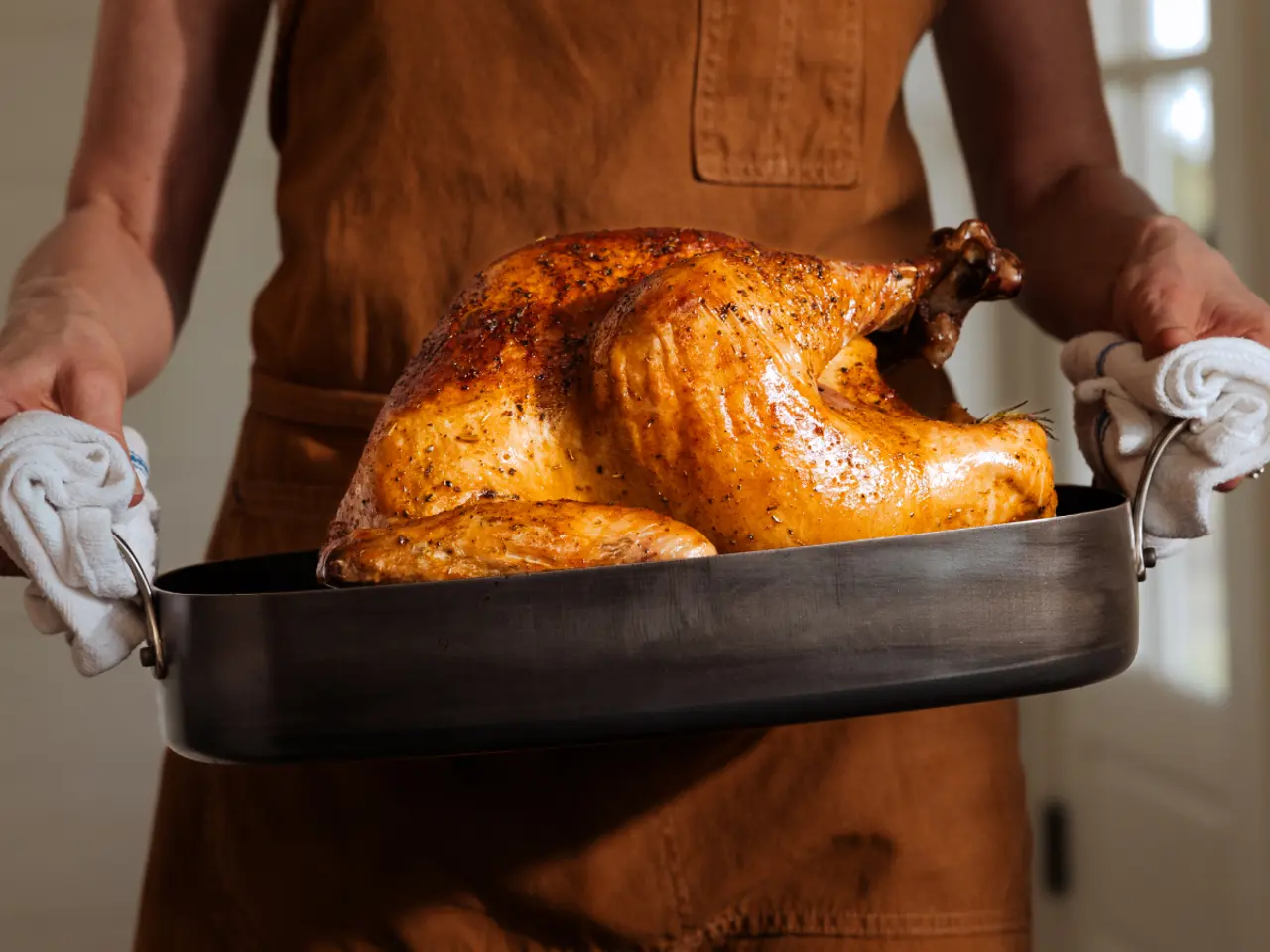 Person in an apron holding a roasted turkey in a pan with oven gloves.