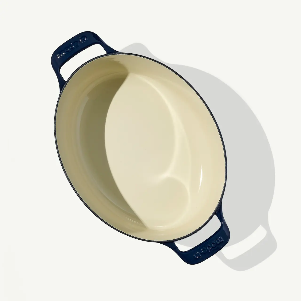 A cream-colored oval baking dish with dark blue handles rests on a light surface casting a soft shadow.