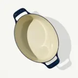 A cream-colored oval baking dish with dark blue handles rests on a light surface casting a soft shadow.