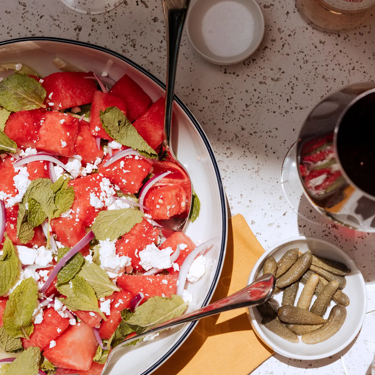 A vibrant salad featuring watermelon, crumbled feta cheese, and fresh mint, accompanied by pickles and a glass of liquid on a textured surface.