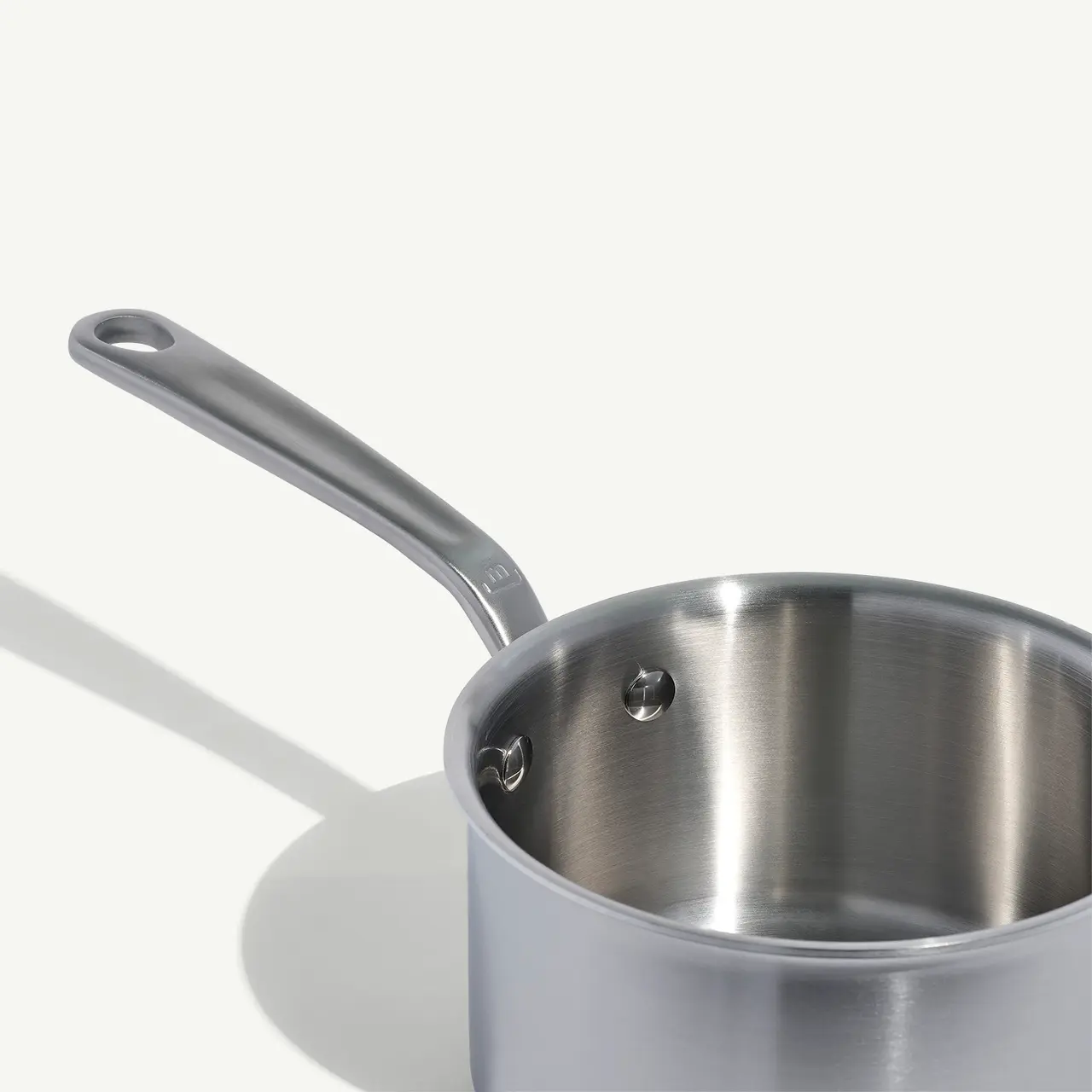 A stainless steel saucepan with a long handle is positioned against a light gray background, casting a soft shadow.
