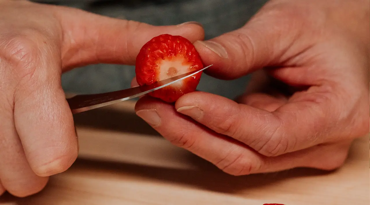 Hands slice a strawberry on a wooden board with a sharp knife.