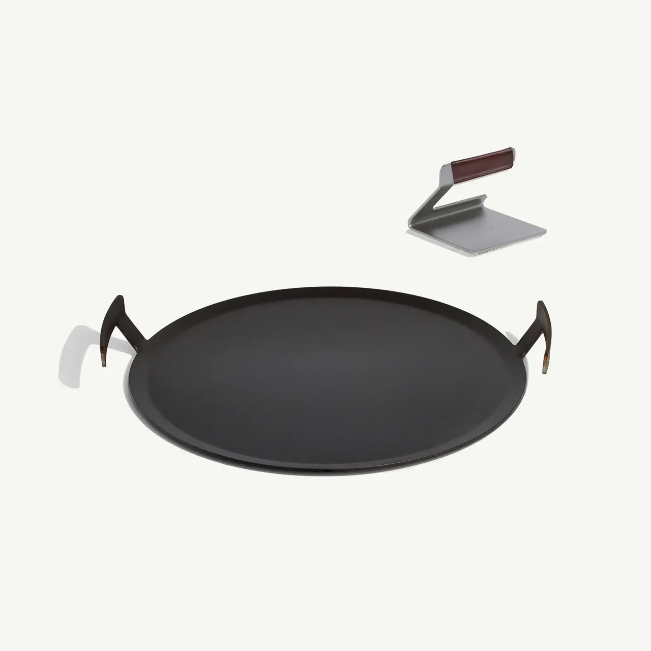 A large black pan with handles is depicted floating above a surface, casting a shadow, accompanied by a metallic spatula with a brown handle leaning against a white holder.