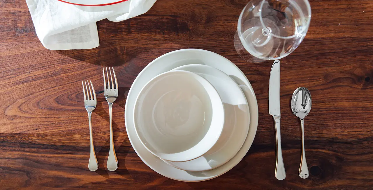 A formal table setting with stacked white plates, a pair of forks to the left, a knife and spoon to the right, and an upside-down wine glass on a wooden table.