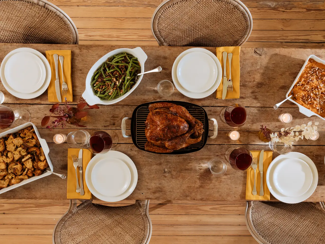 A top-down view of a rustic dining table set for a meal with a roasted chicken centerpiece, surrounded by various side dishes, cutlery, napkins, and glasses of wine.