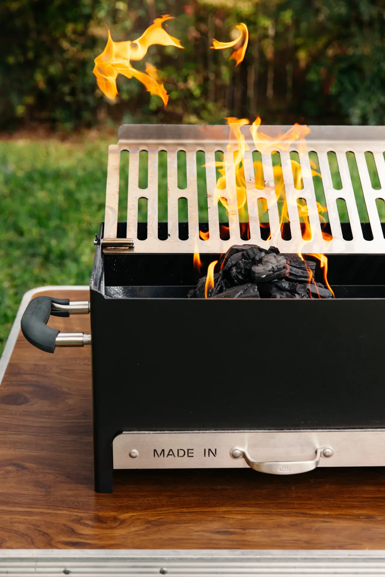 A barbecue grill with flames rising from hot coals is placed on a wooden surface in an outdoor setting.
