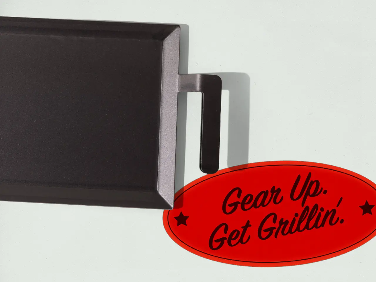 A close-up view of a black gadget with a silver handle attached to a red oval sign that reads "Gear Up. Get Grillin'."