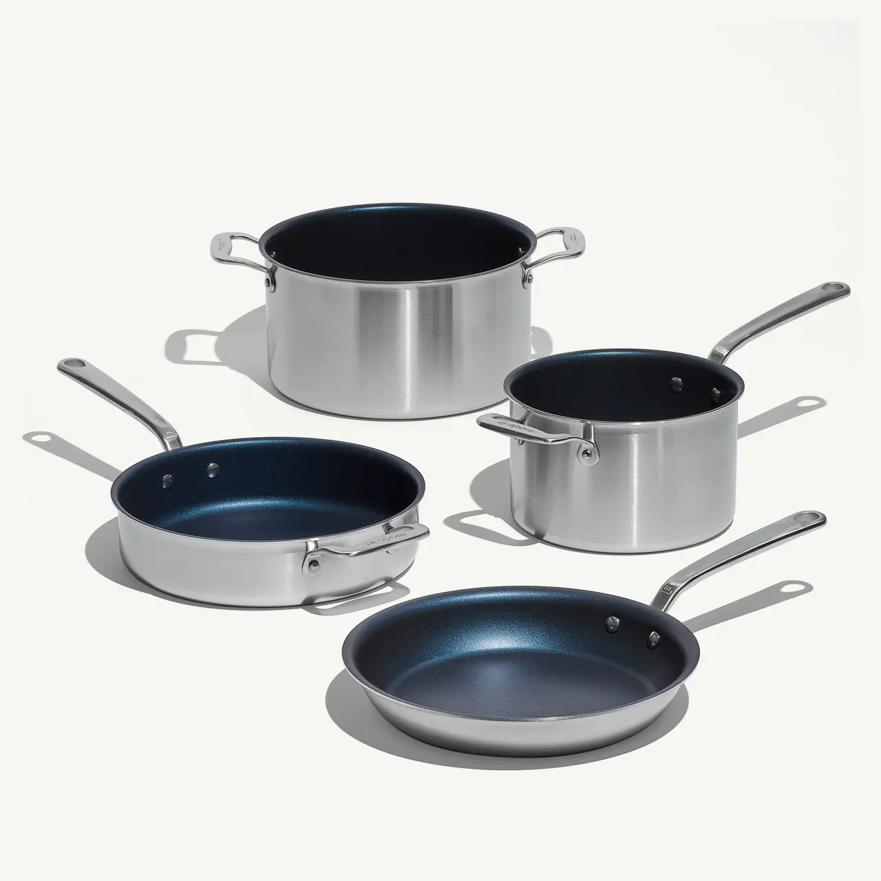 A set of stainless steel cookware, including two frying pans and two pots with blue non-stick interiors, is arranged on a light background.