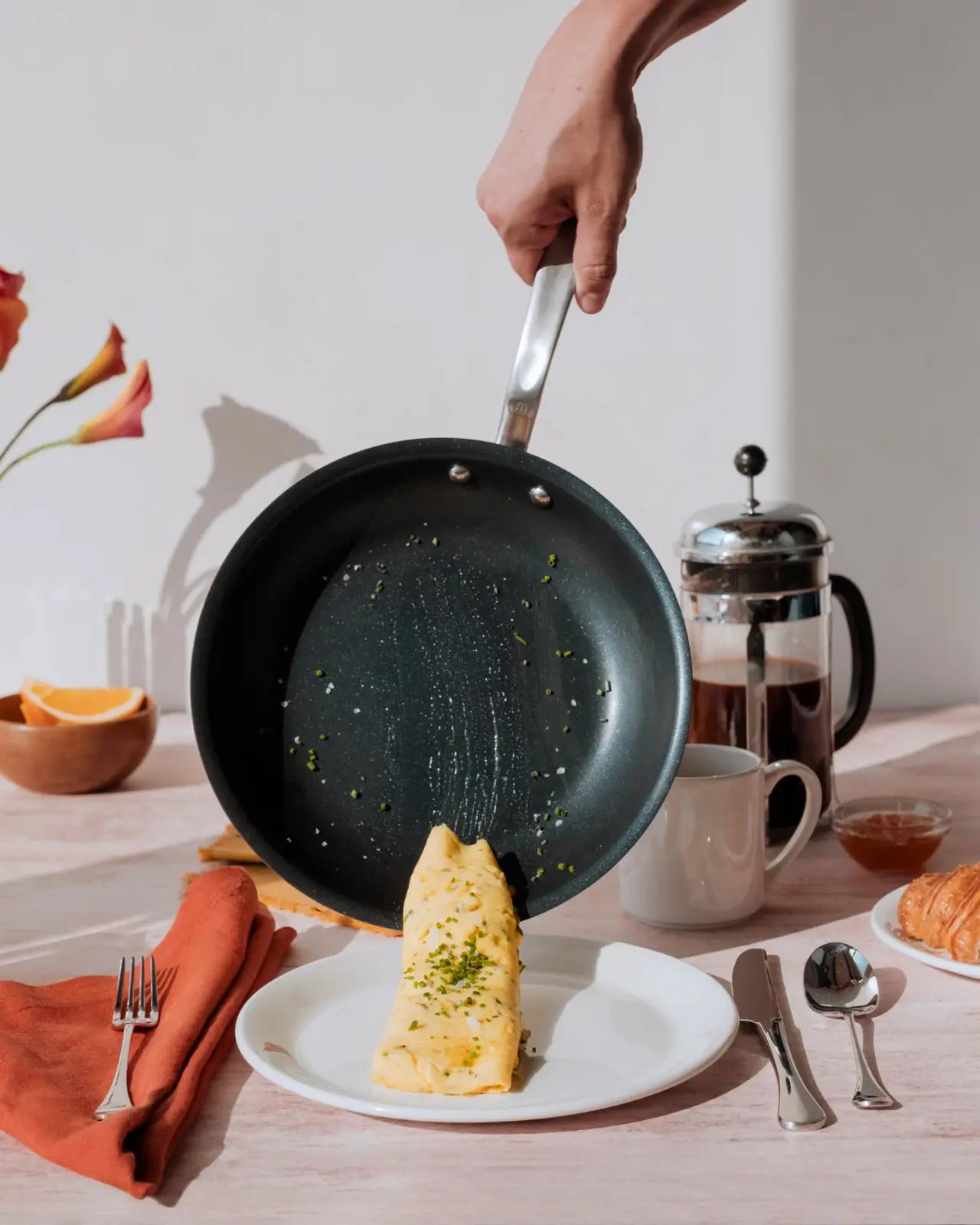 A hand is flipping an omelette from a frying pan onto a plate set for breakfast with a French press and a croissant on the table.