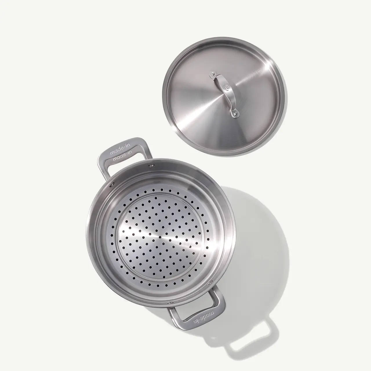 A stainless steel pot with a colander insert is placed next to its lid on a light surface.