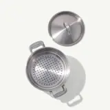 A stainless steel pot with a colander insert is placed next to its lid on a light surface.