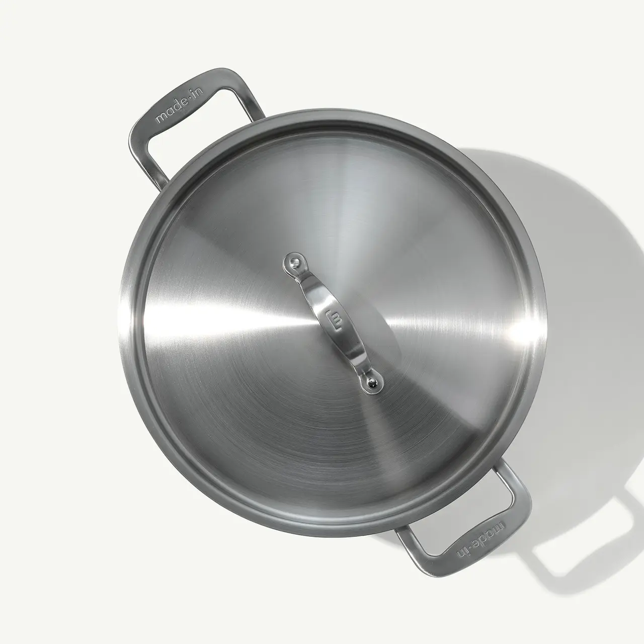 A stainless steel pan with a lid is shown from above, casting a soft shadow on a light surface.