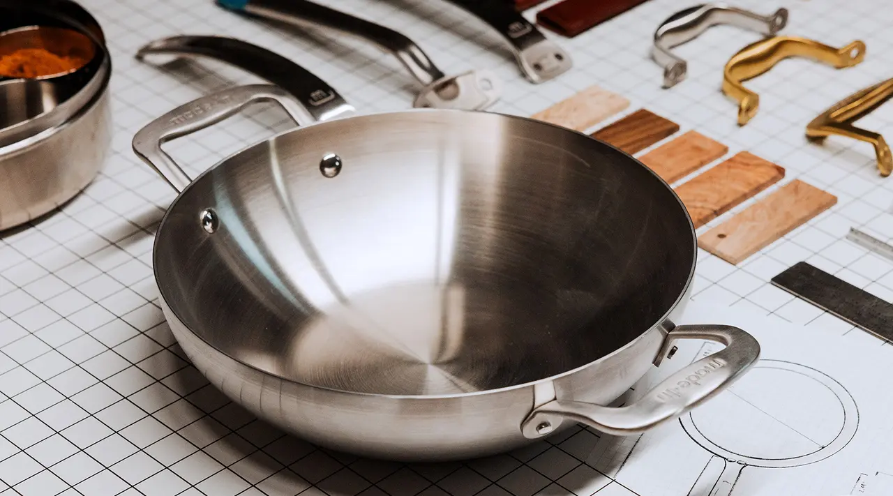 A stainless steel frying pan is centered on a grid paper with various kitchen utensils and handles scattered around it.