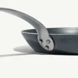 A close-up view of a black frying pan's handle attached with rivets, set against a light background.