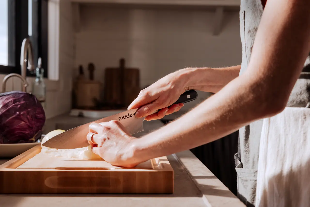A person is slicing an onion on a wooden cutting board in a sunlit kitchen.