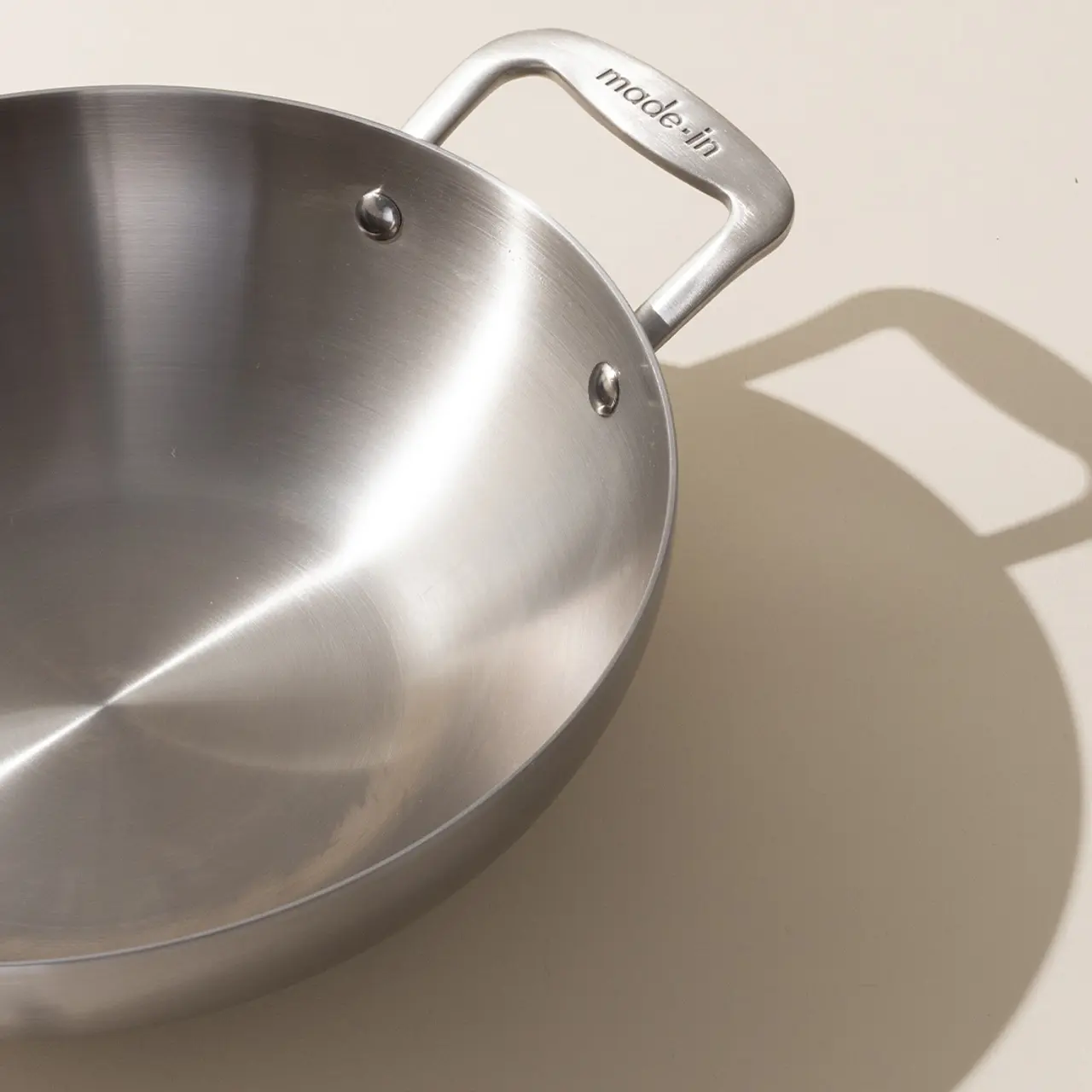 A stainless steel frying pan with the brand name 'made in' on the handle, casting a shadow on a light surface.