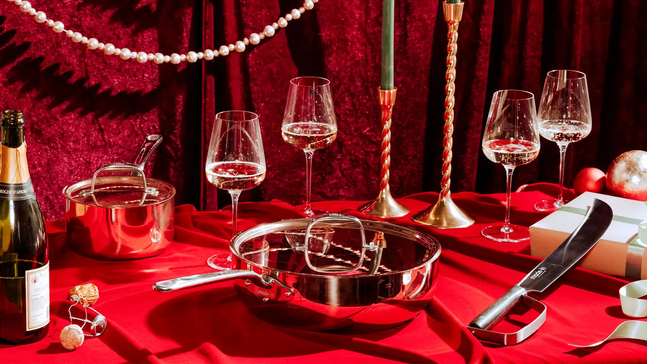 An elegant festive table setting featuring a bottle of champagne, various wine glasses, stainless steel cookware, and red and gold decorations on a red tablecloth.