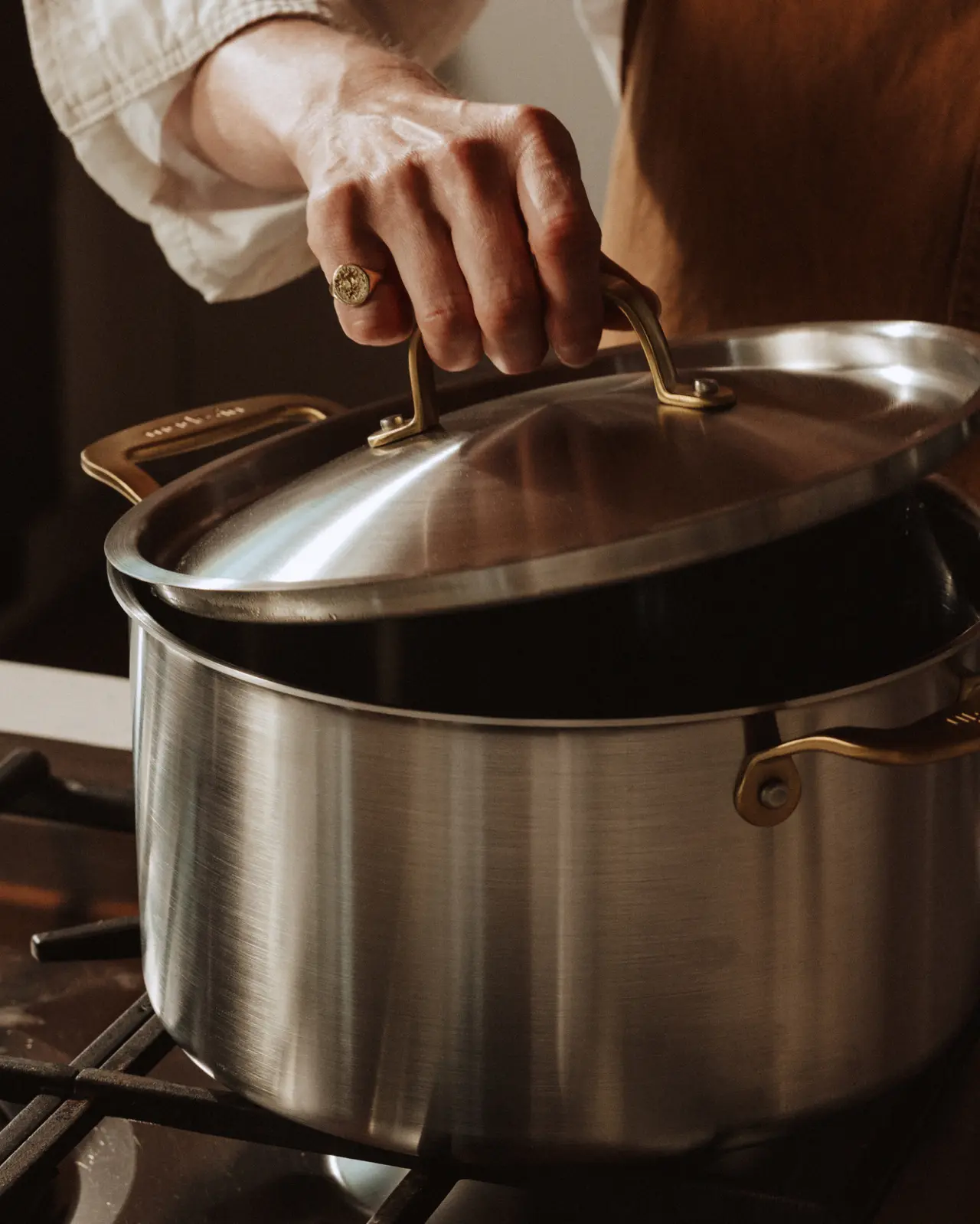 A person's hand with a ring is lifting the lid off a stainless steel pot on a gas stove.