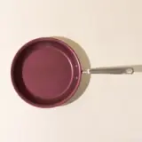 A top-down view of a burgundy-colored frying pan with a silver handle on a beige surface.