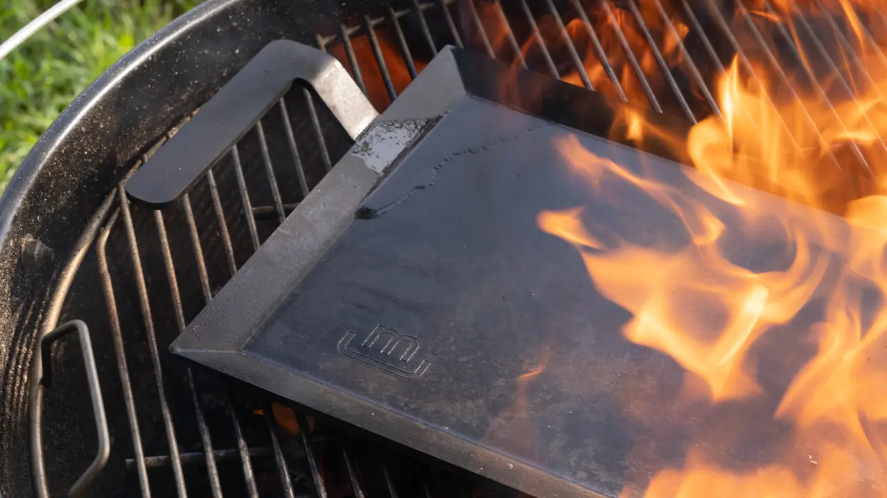 Flames lick the edge of a closed grill topper on a barbecue with a vibrant fire underneath.