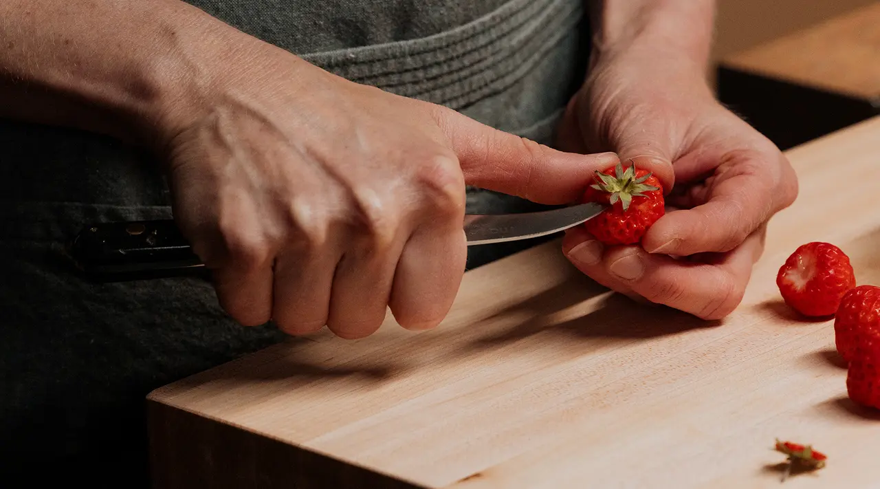 Close-up of hands using a knife to hull a strawberry on a wooden cutting board.