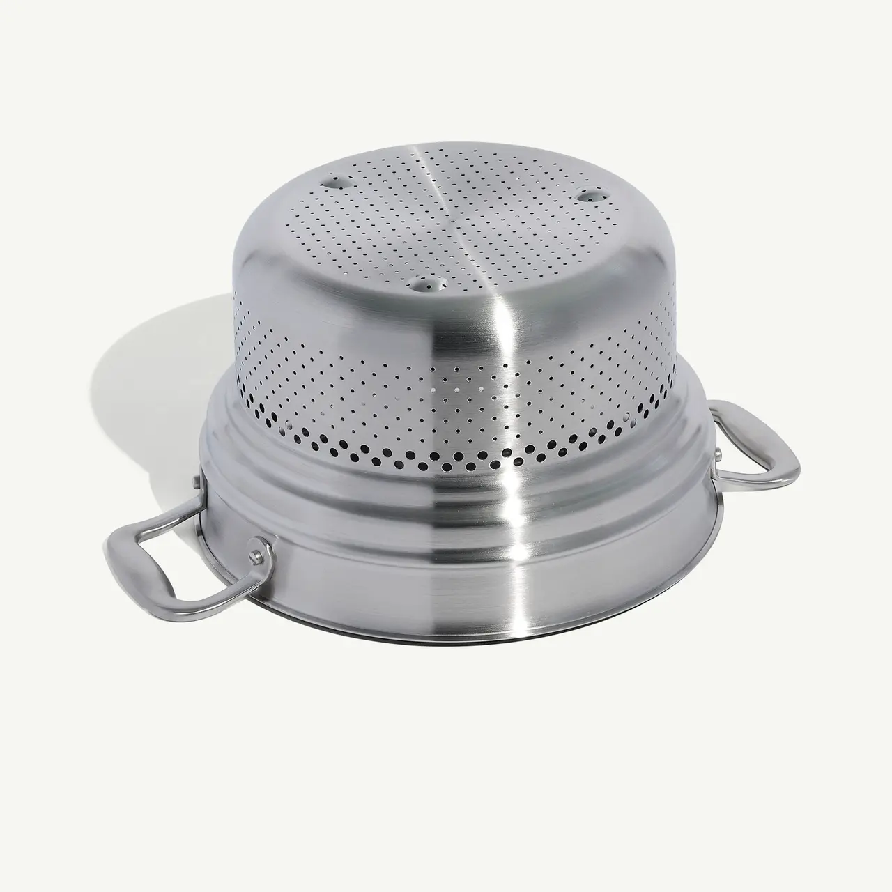A stainless steel food steamer insert with perforated top and side handles rests on a light background.
