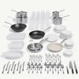 A collection of kitchenware including pots, frying pans, plates, cutlery, and glass cups arranged neatly on a light background.