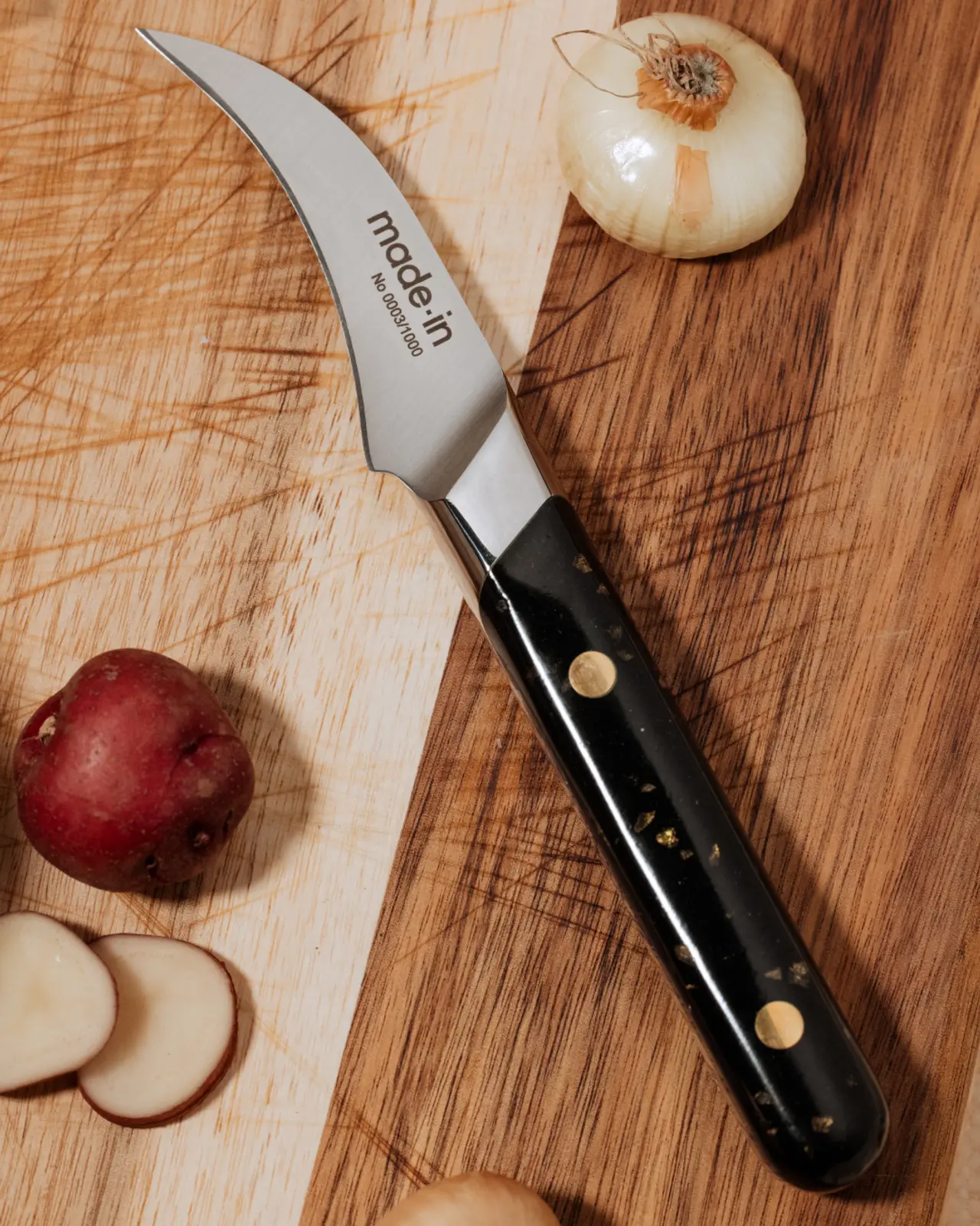 A sharp kitchen knife lies on a wooden cutting board next to a partly sliced red apple and a whole white onion.