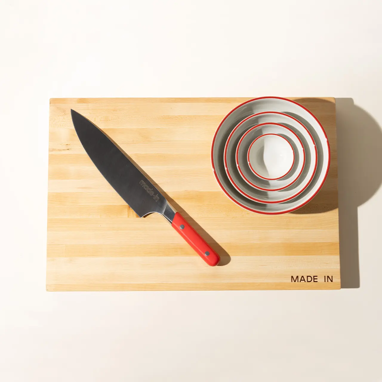 A chef's knife lies next to a stack of nested bowls on a wooden cutting board with "MADE IN" text.
