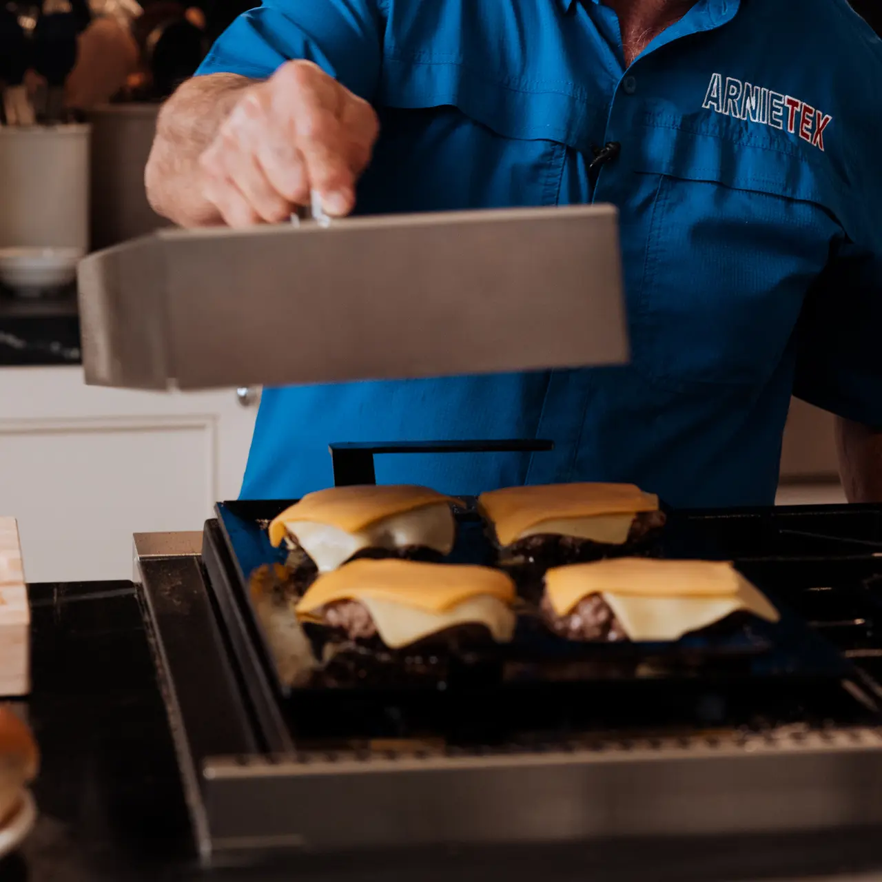 A person in a blue shirt cooks burgers with cheese on a grill using a large metal spatula.