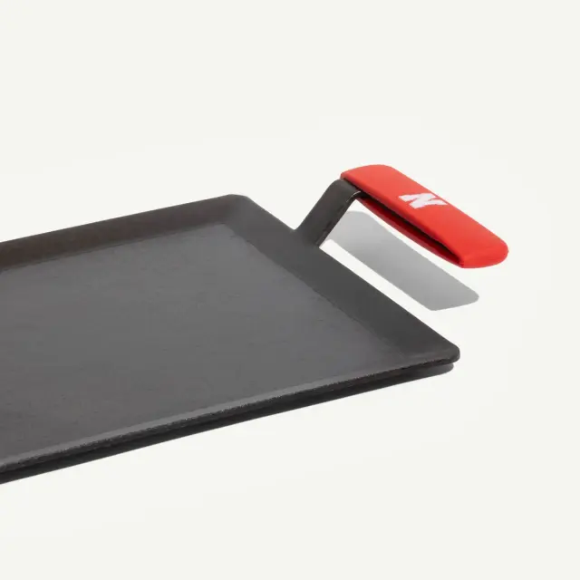 A black rectangular griddle with a red handle and a metal spatula attachment.