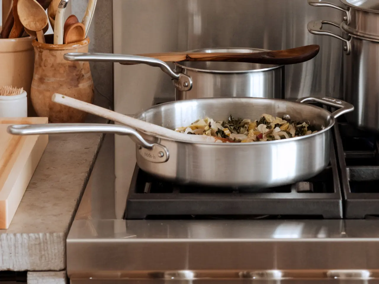 A stainless steel sauté pan with wooden-handled utensils, containing food, sits on a stove next to other kitchen pots and wooden cooking tools.