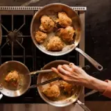 Golden-brown chicken pieces are frying in two sauté pans on a stovetop, with a hand maneuvering a spatula to check their doneness.