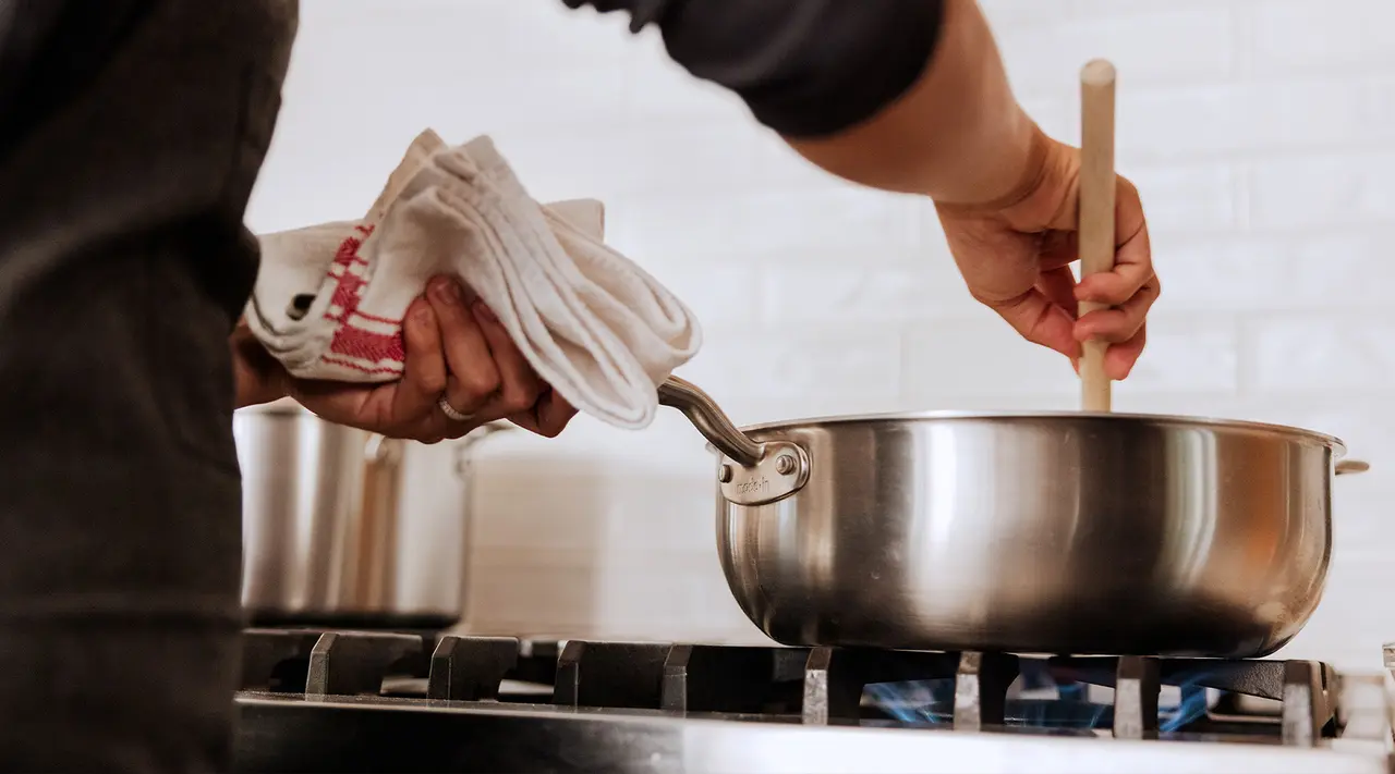 A person is cooking, stirring a pot on a stove while holding a kitchen towel.