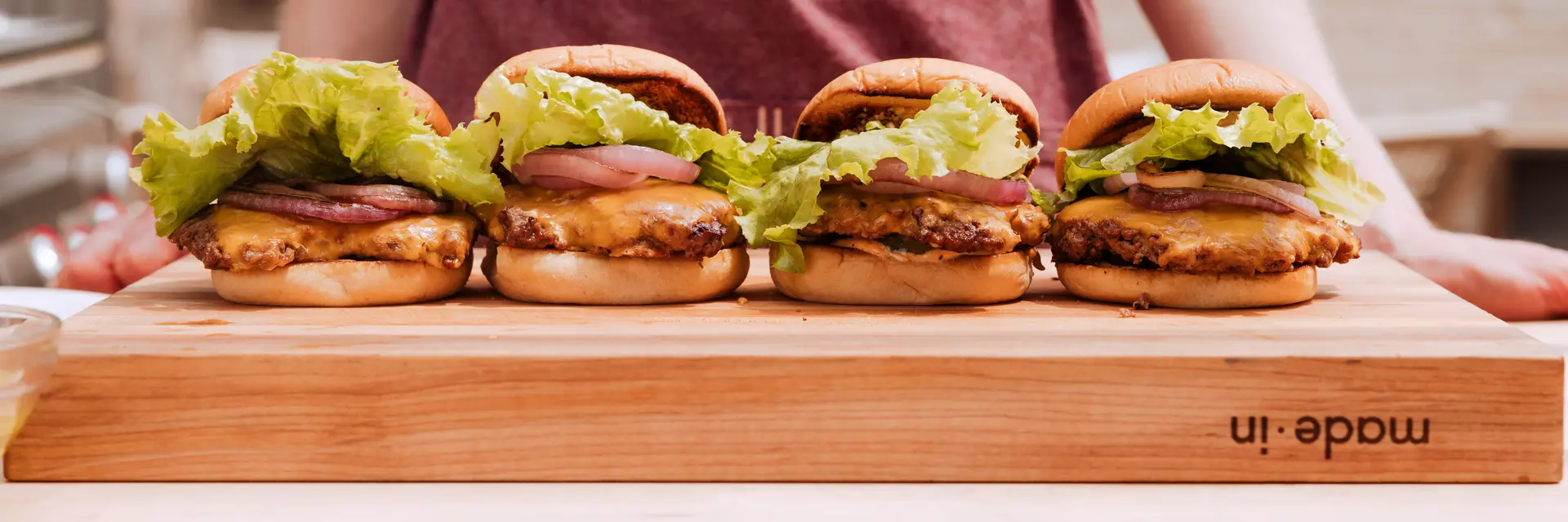 A person is holding a wooden board with three appetizing burgers lined up on it.