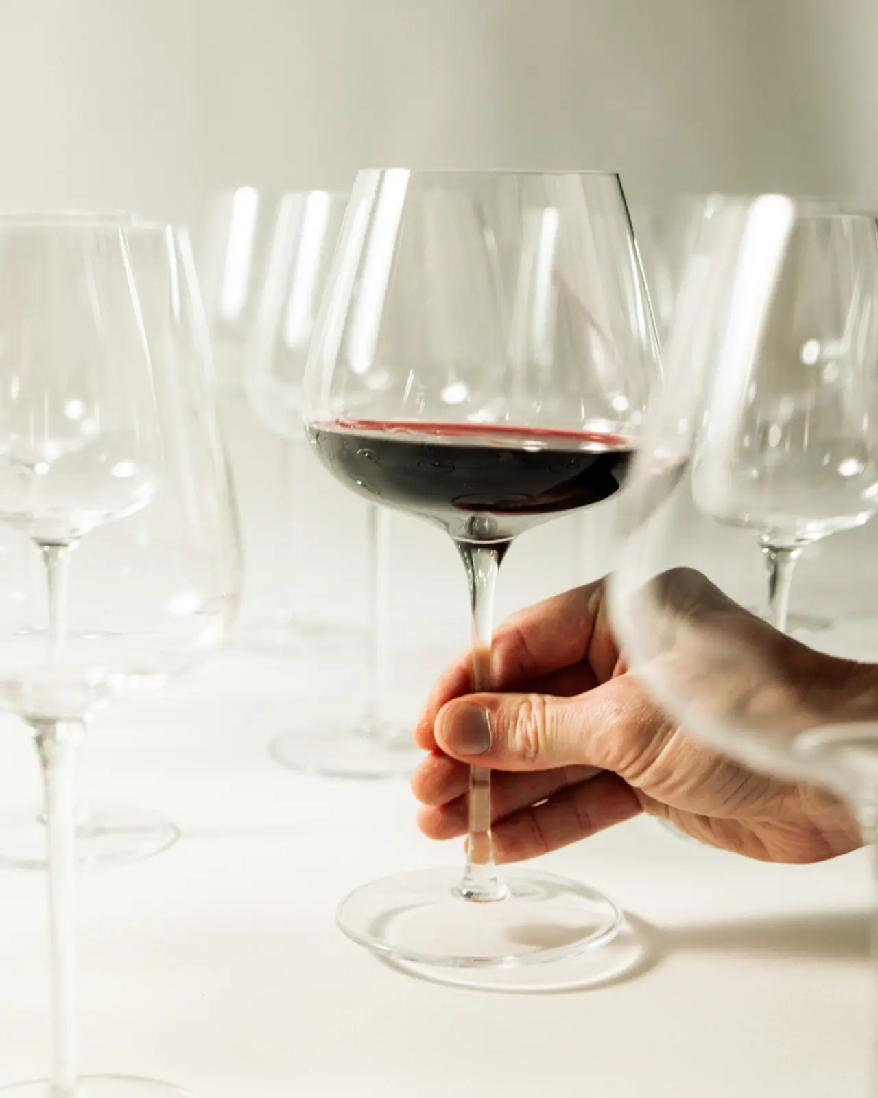 A hand holds a stem of a wine glass filled with red wine amidst several empty glasses on a white background.
