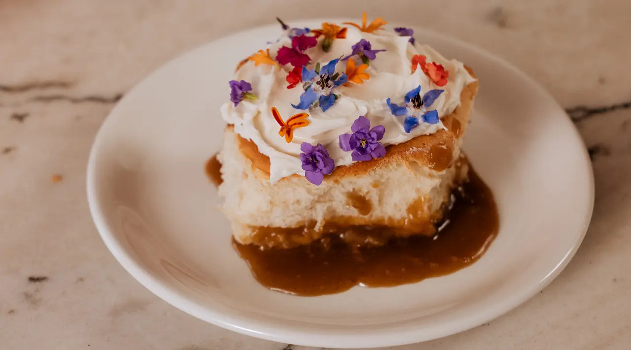 A piece of cake topped with white frosting and colorful edible flowers sits on a white plate with a drizzle of caramel sauce.