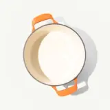 An empty orange-handled enameled pot casts a soft shadow on a pale background.