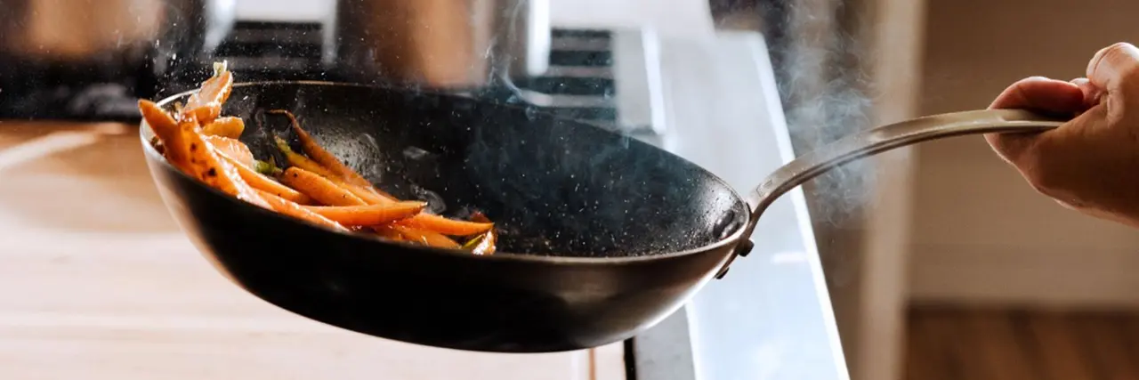 A person is tossing carrots in a frying pan over a kitchen stove.